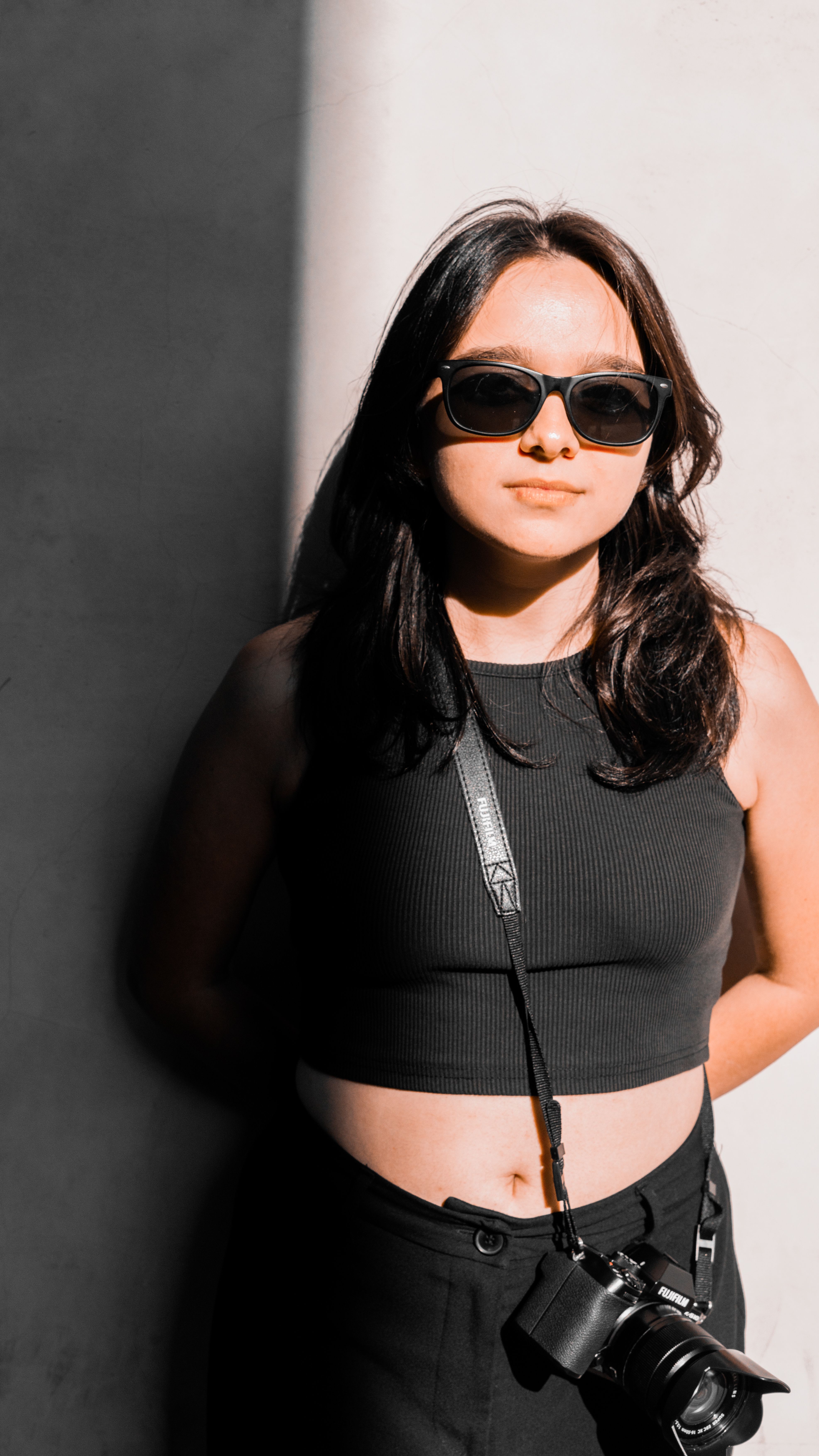 Portrait of a fashionable woman wearing sunglasses and a black crop top, standing confidently against a shaded wall.