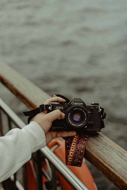 Free Close-up of a hand holding a vintage camera against a wooden railing overlooking water. Stock Photo
