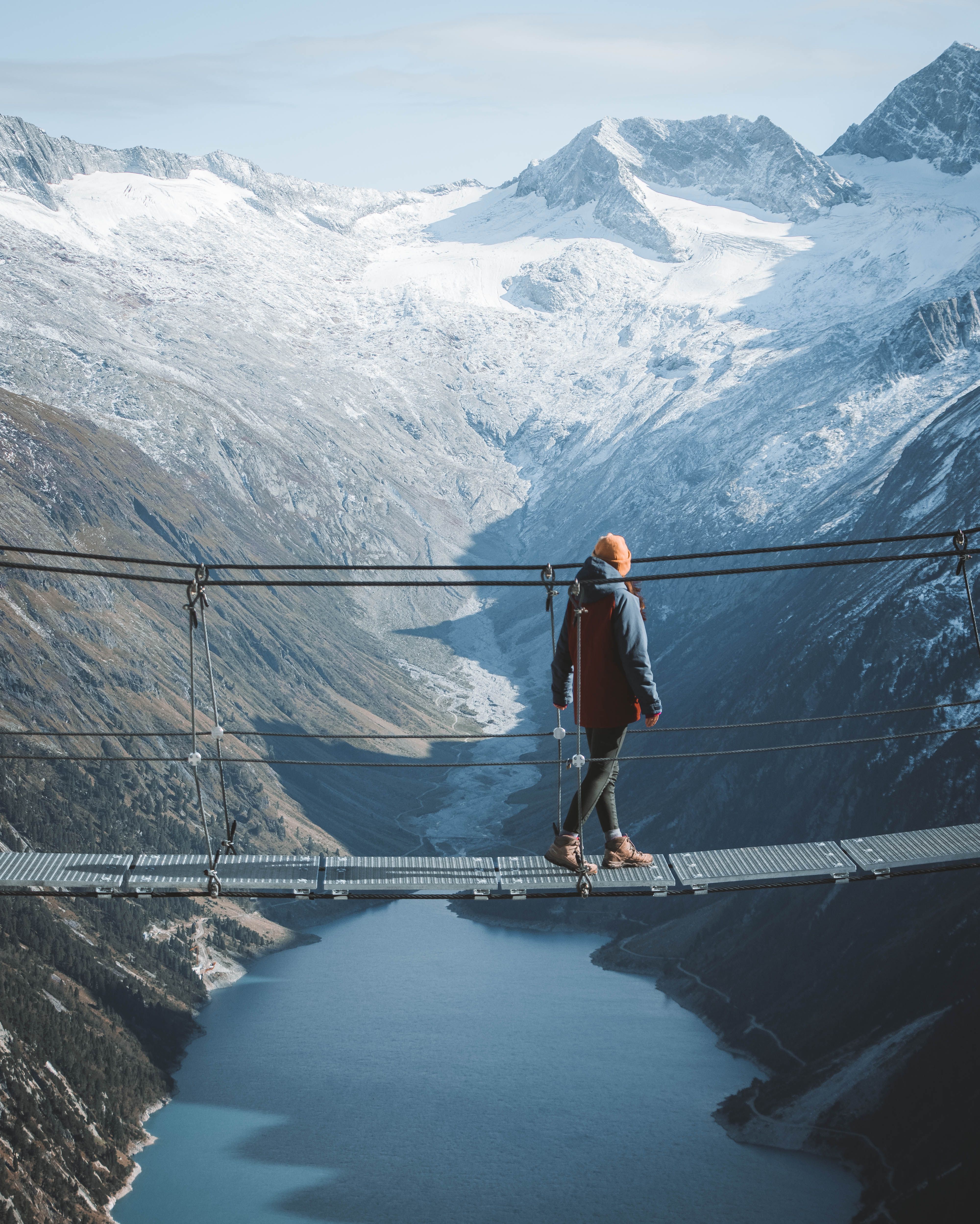 A person walks on a suspension bridge amidst snowy alpine mountains.