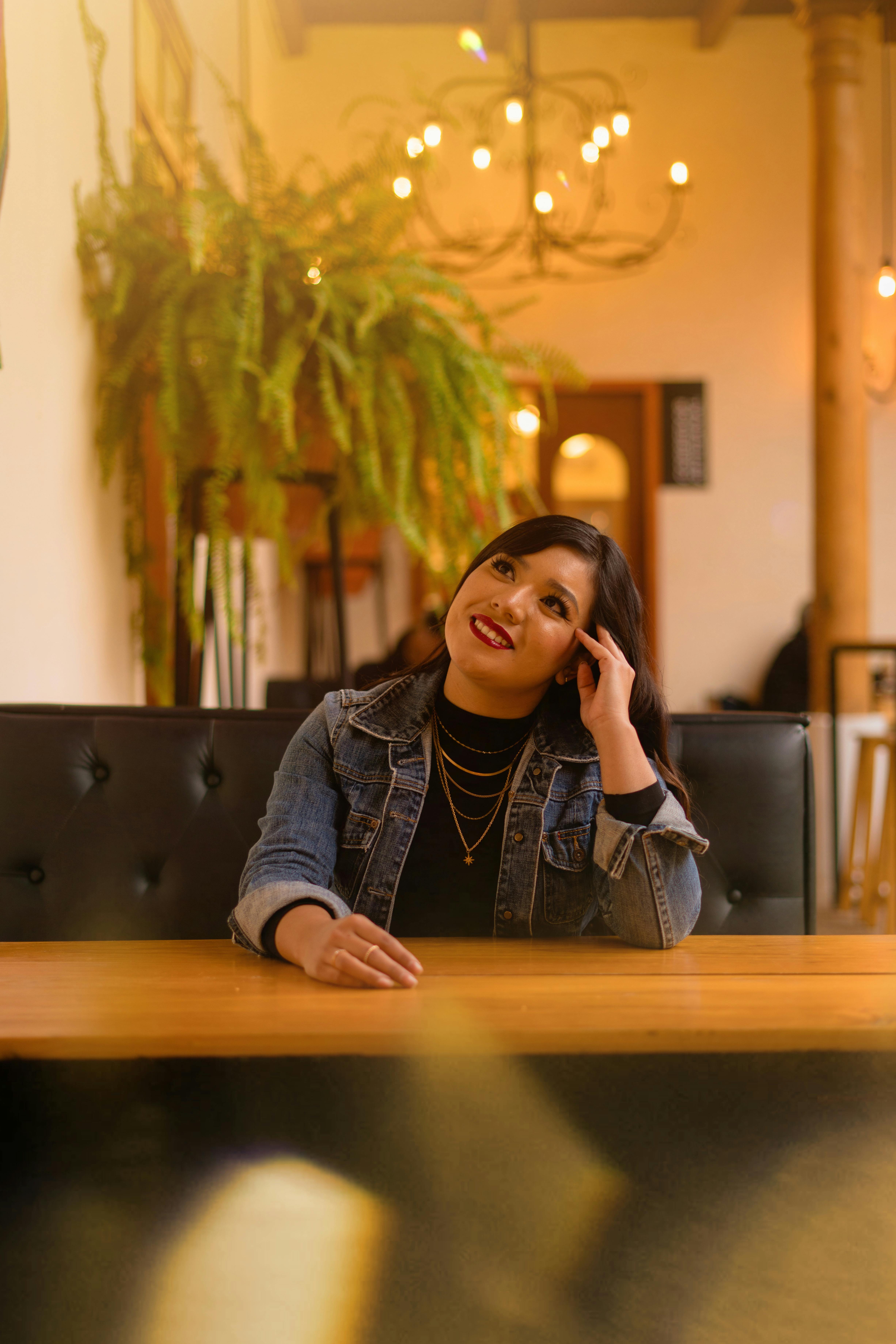 Portrait of a woman in a denim jacket at a warmly lit restaurant table, smiling.
