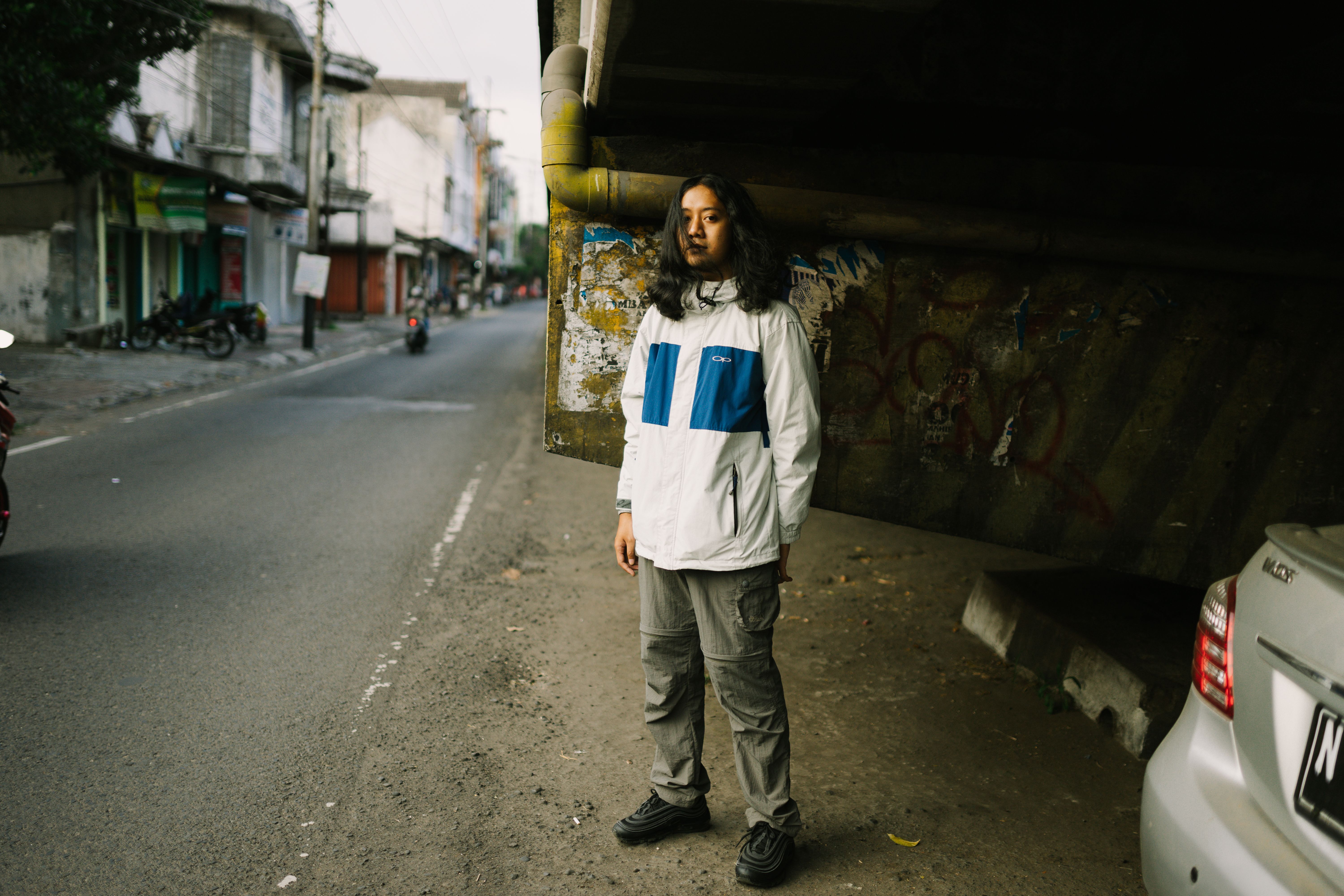 A person in a stylish jacket stands confidently on a city street under a bridge.