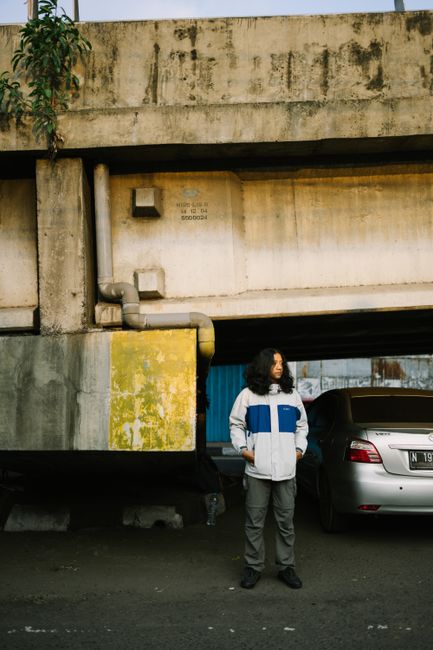 Person in a white jacket stands under bridge beside parked car, urban setting.