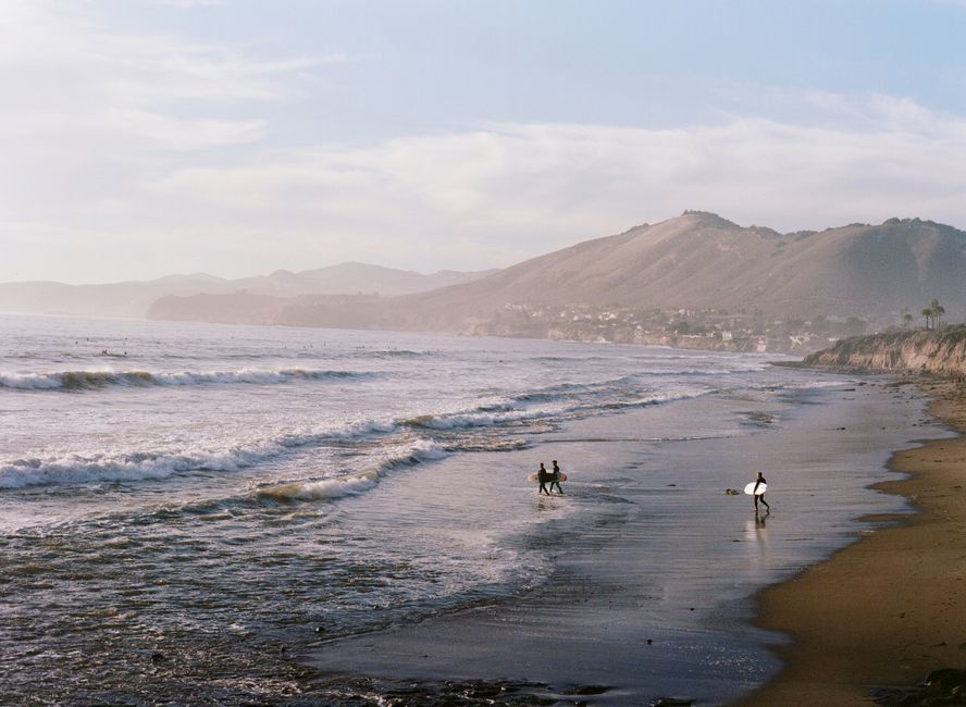 Surfers walk along the serene Pismo Beach coastline during a summer day with waves crashing softly.
