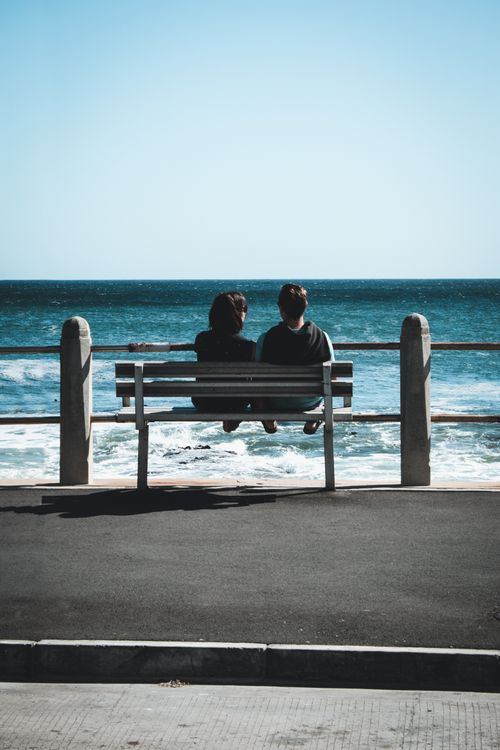 Free A couple enjoys a peaceful moment on a bench by the sea, under a clear blue sky. Stock Photo
