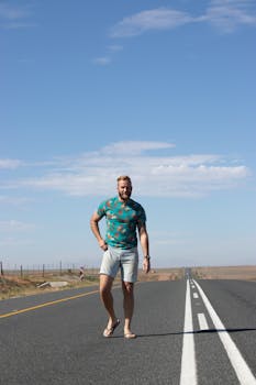 Man in denim shorts and printed shirt standing on open road under blue sky.