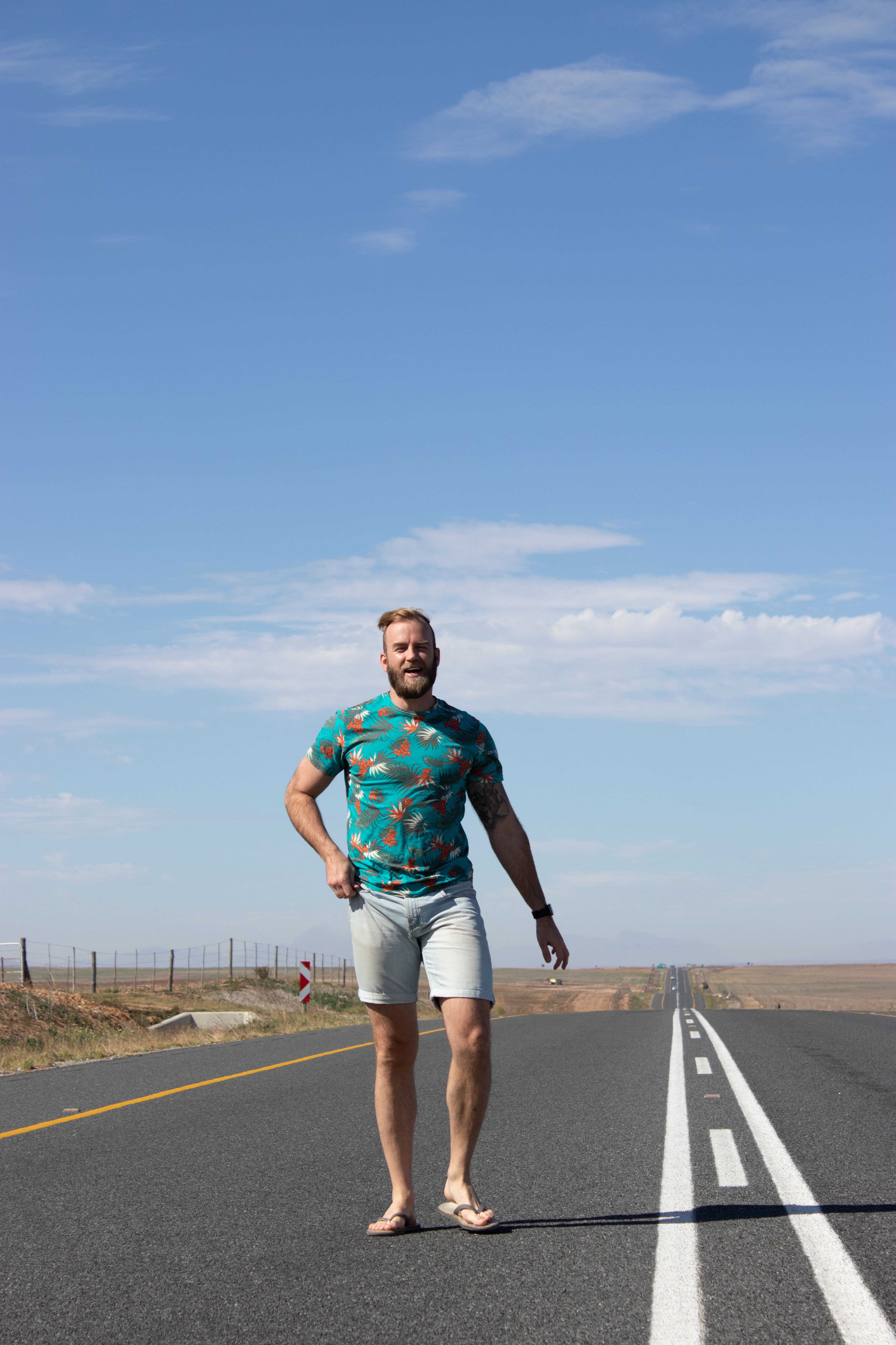 A man with a beard and printed shirt walks down a highway under a bright blue sky with clouds.