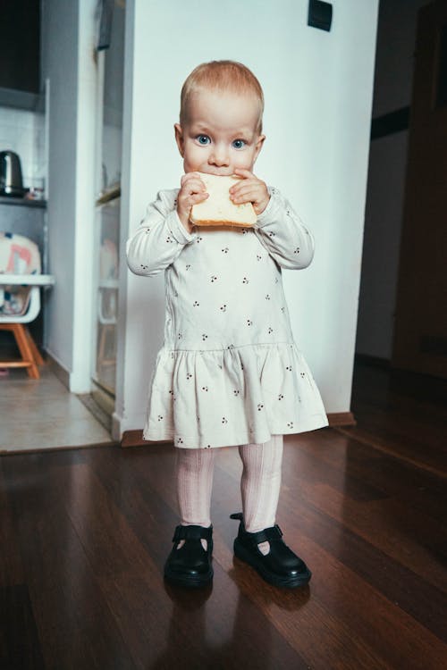 A Young Girl in White Dress Standing on a Wooden Floor while Eating Bread