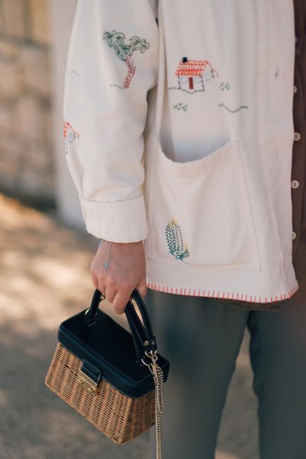 Close-up of a person holding a chic handbag, wearing an embroidered overshirt in natural light.