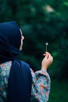 A woman in a hijab holds a dandelion, gently blowing seeds away in a serene outdoor setting.