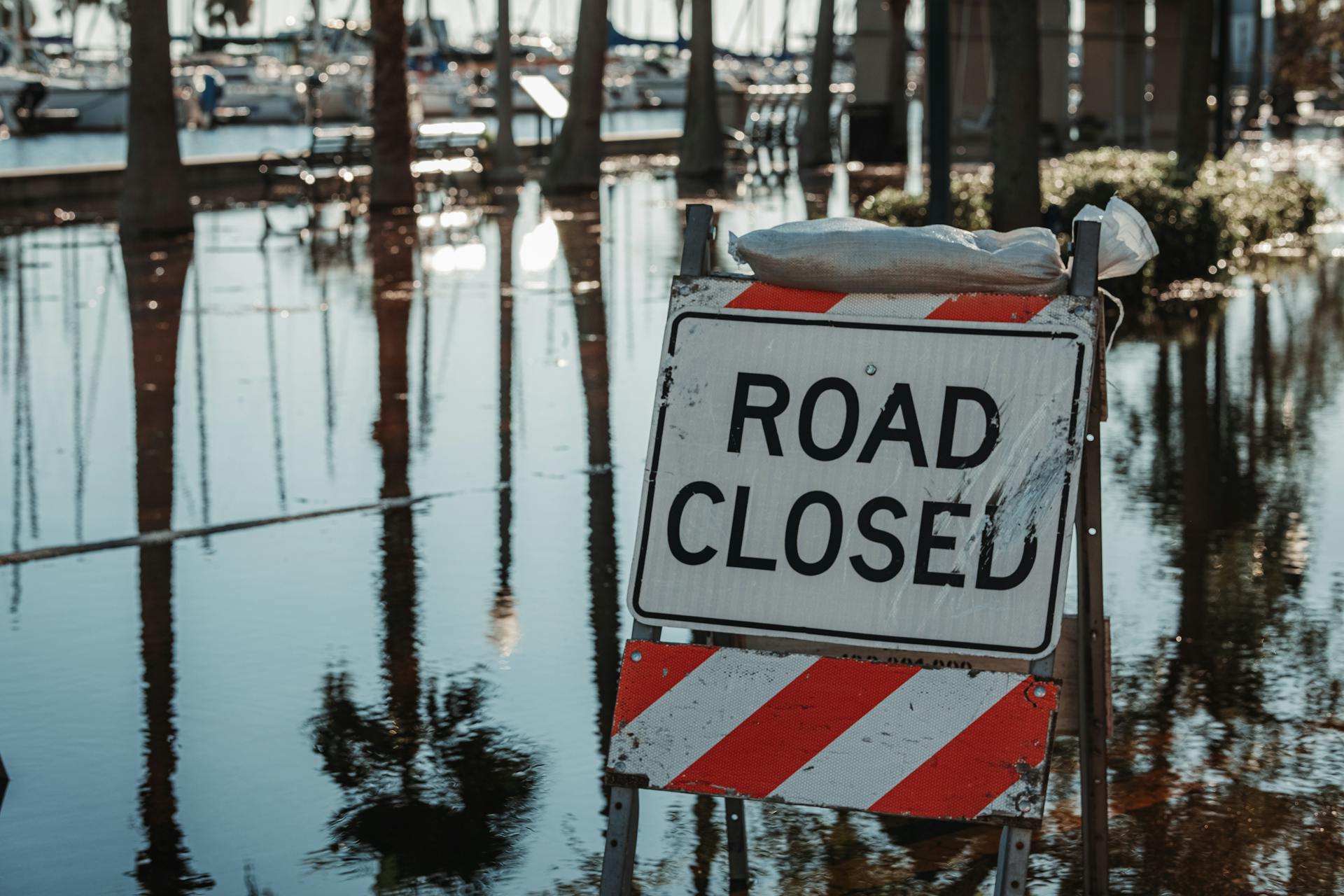 Road closed sign amidst flooded street, reflecting calm water and trees.