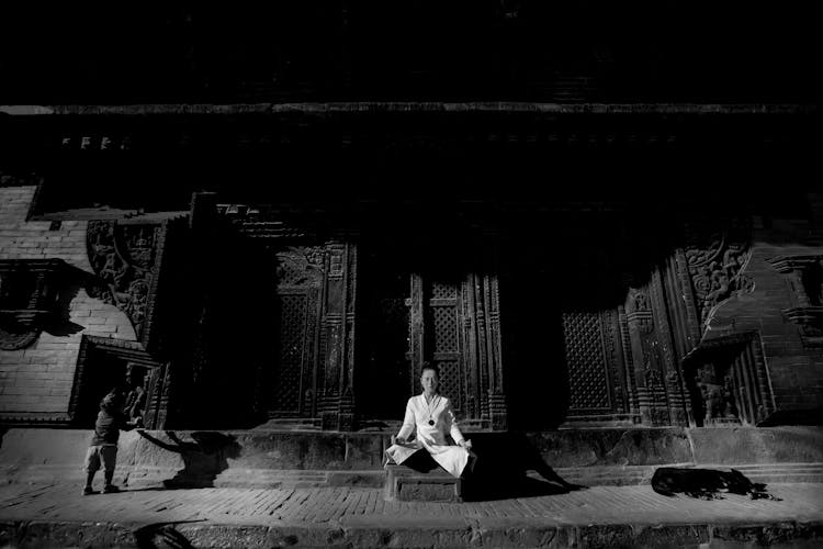 Woman Wearing White Clothing Meditating In Front Of A Dark Wooden Ornamental Architecture