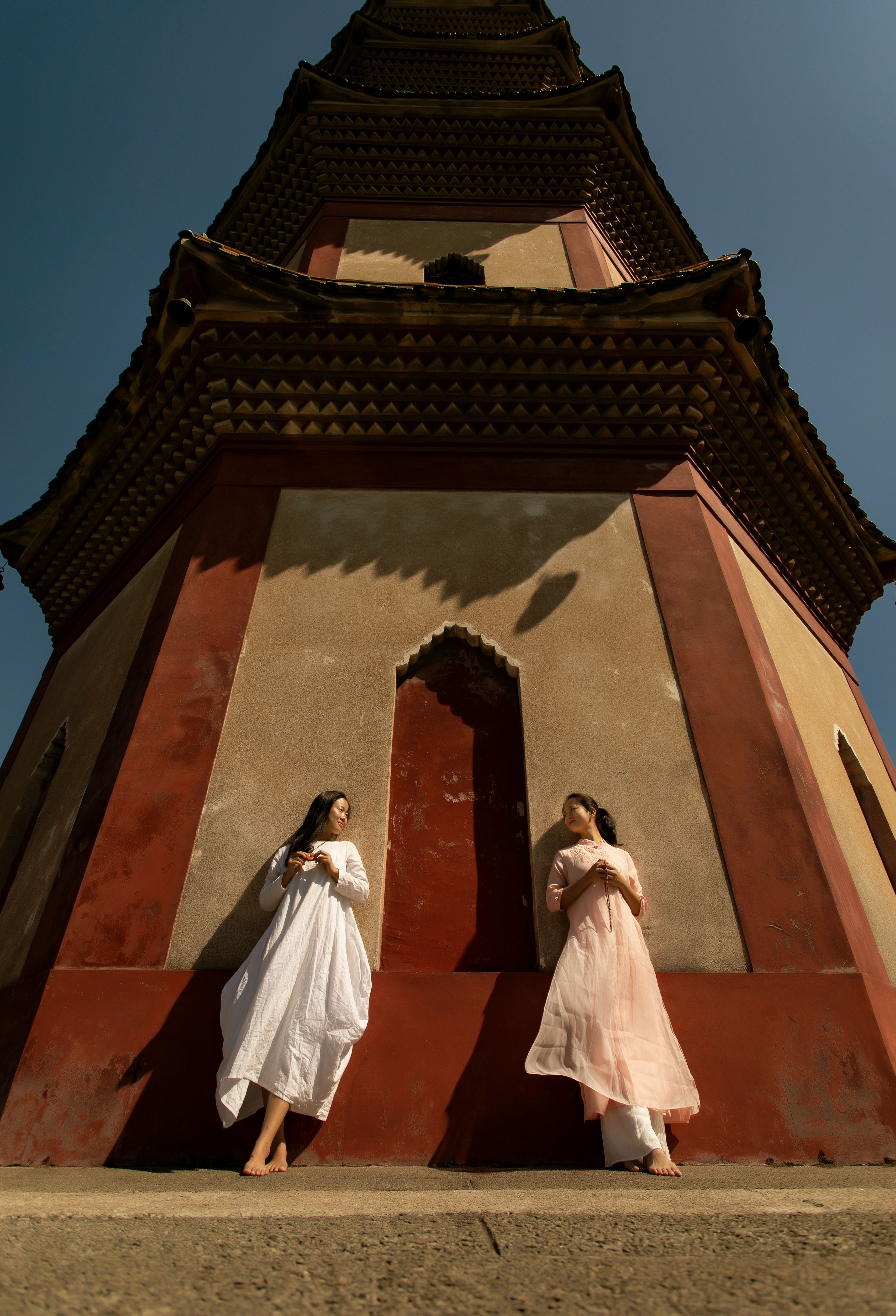Two women in elegant dresses pose by a towering Asian architectural structure.