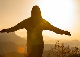 A woman stands with arms open, embracing the sunrise in a mountainous landscape, creating a tranquil silhouette.