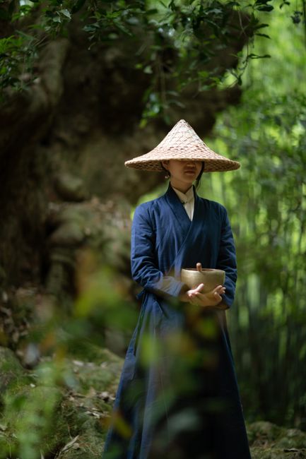 Monk wearing conical hat holding a Tibetan singing bowl in peaceful forest setting.