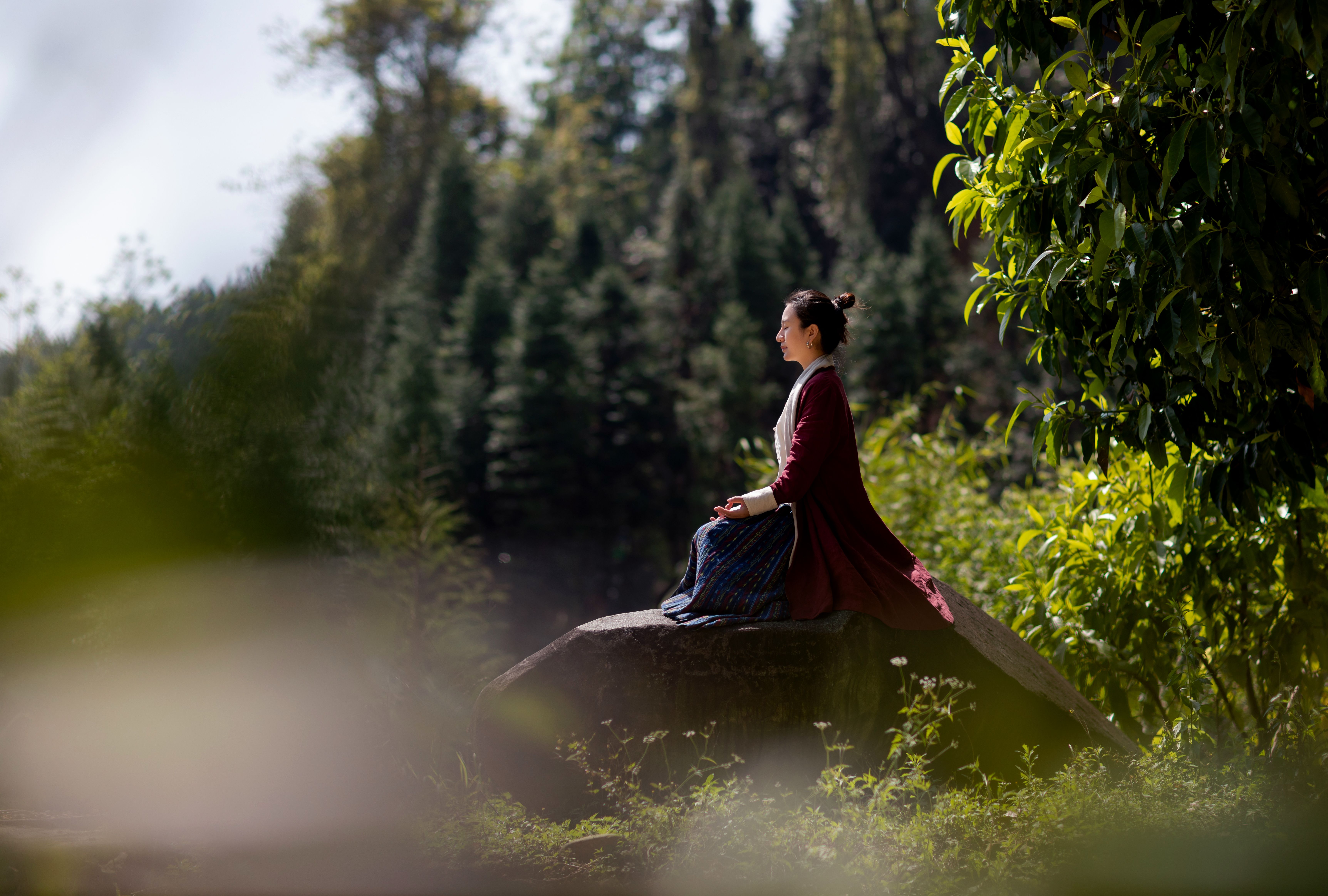 A woman in a traditional costume meditating on a rock amidst nature.