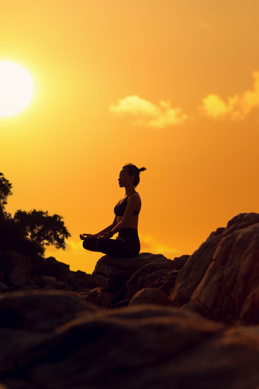 Silhouette of a person sitting in lotus pose on coastal rocks against a golden sunset sky — photo by RAY LEI on Pexels