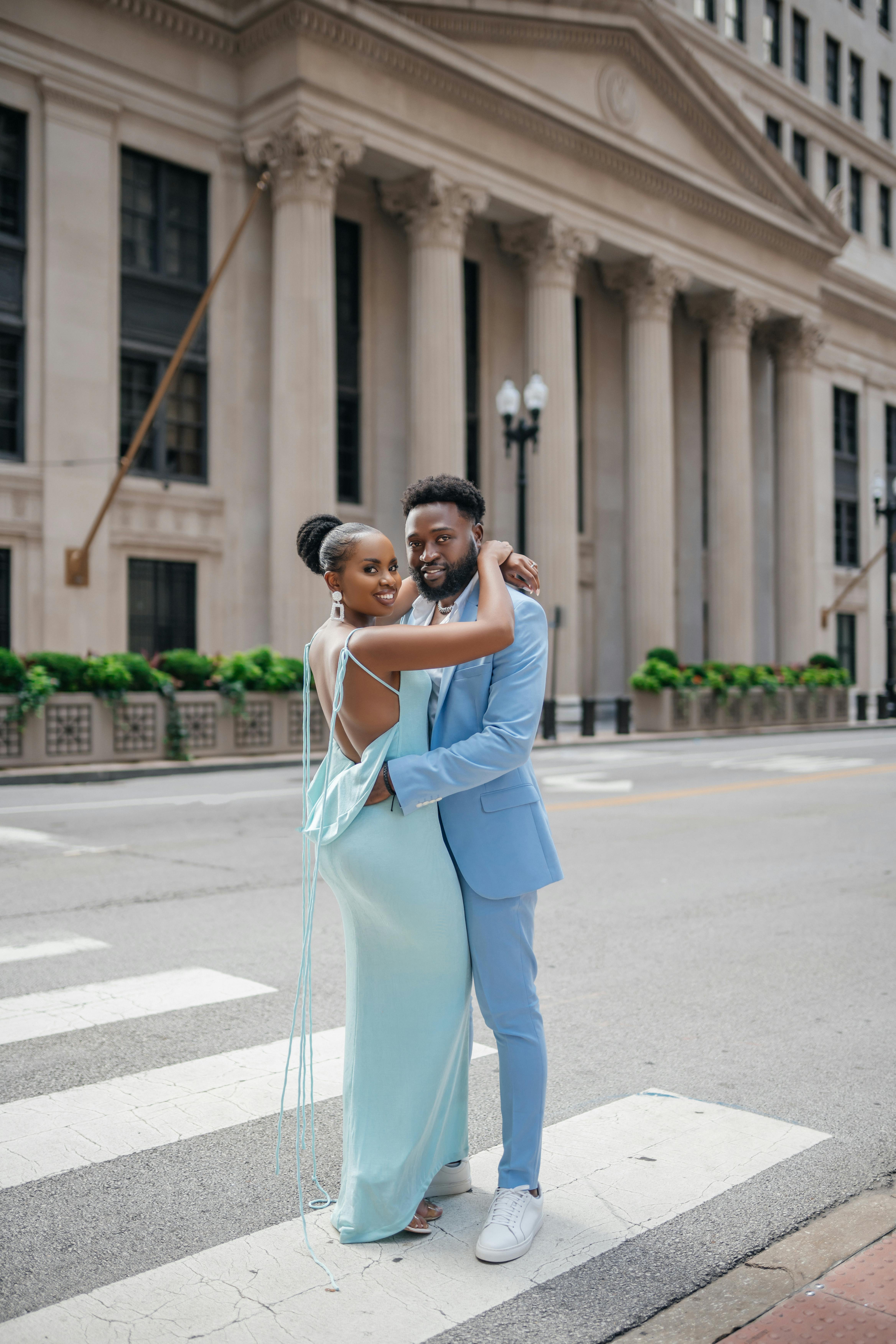 Stylish couple in pastel outfits embracing at a city crosswalk in front of a grand building.