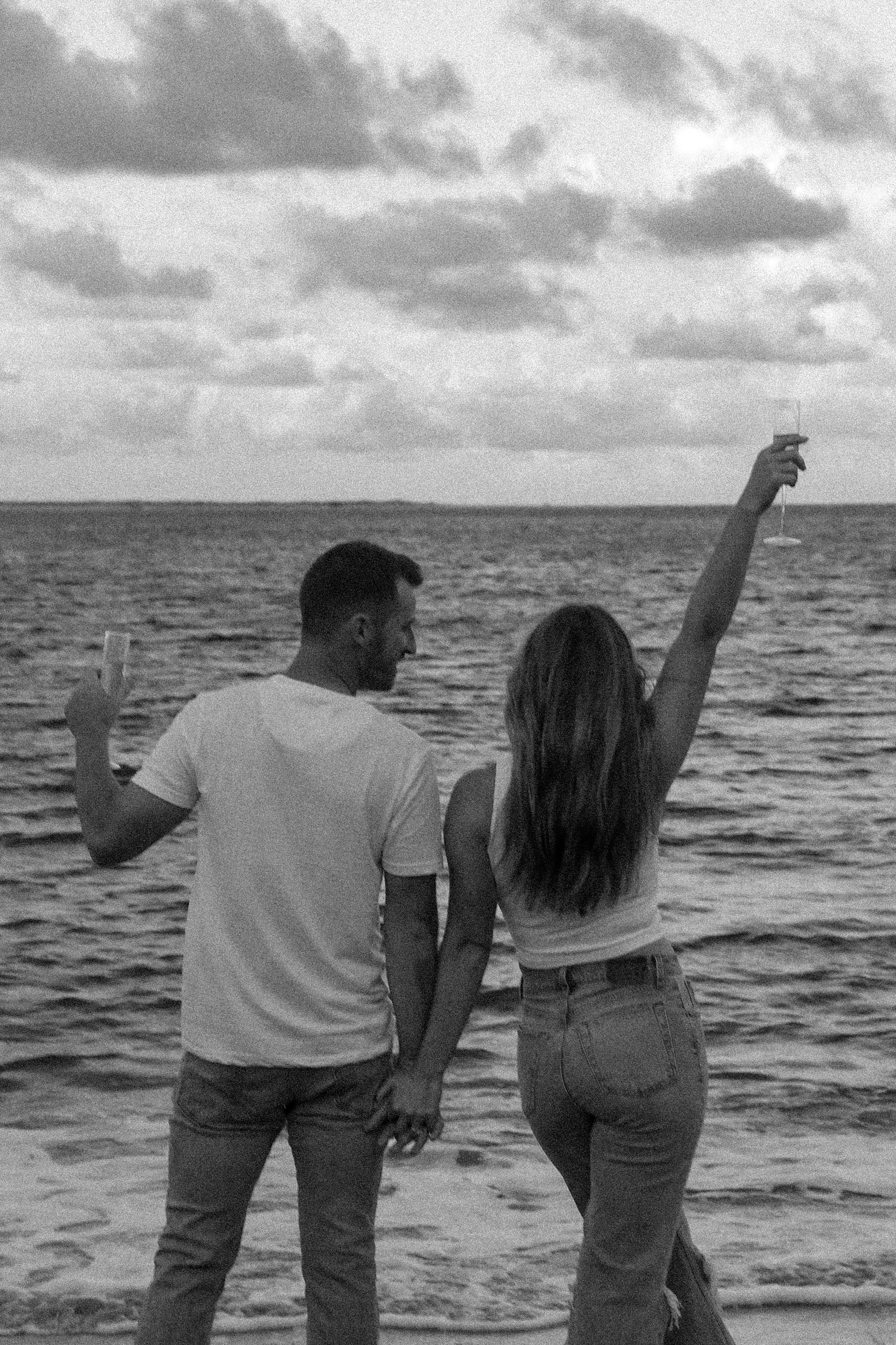 A couple holding hands and toasting with champagne by the ocean, symbolizing love and celebration.