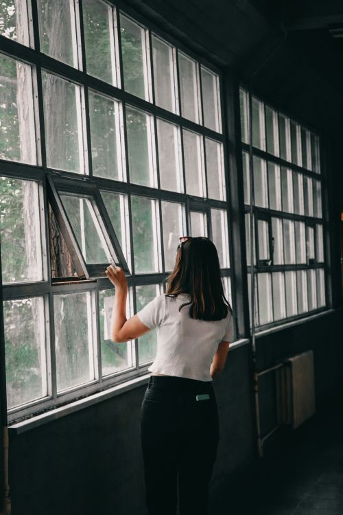 Free Woman in white shirt and black pants opening a large window indoors, showcasing an industrial setting. Stock Photo