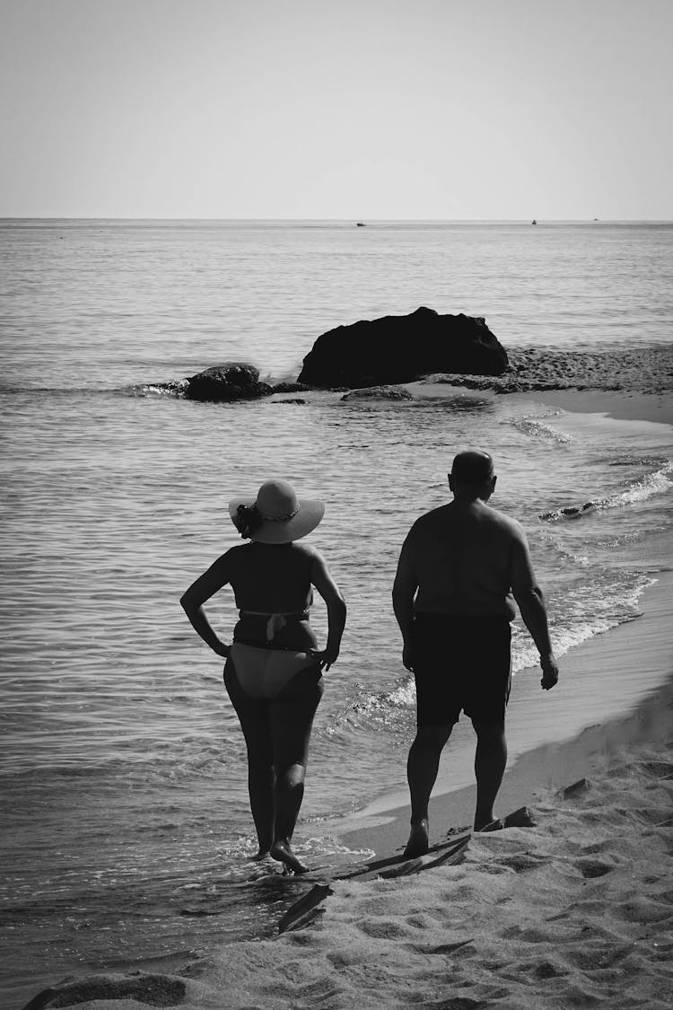 Grayscale Photo Of A Senior Couple In Swimwear Walking On Seashore