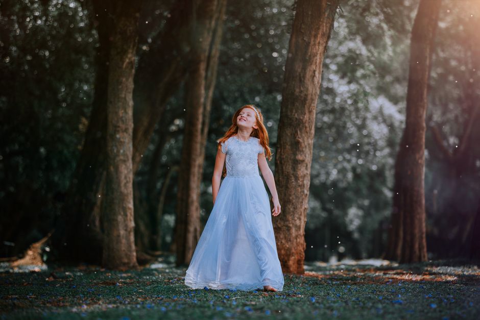 A young girl in a white gown enjoys the magical atmosphere of a sunlit forest.
