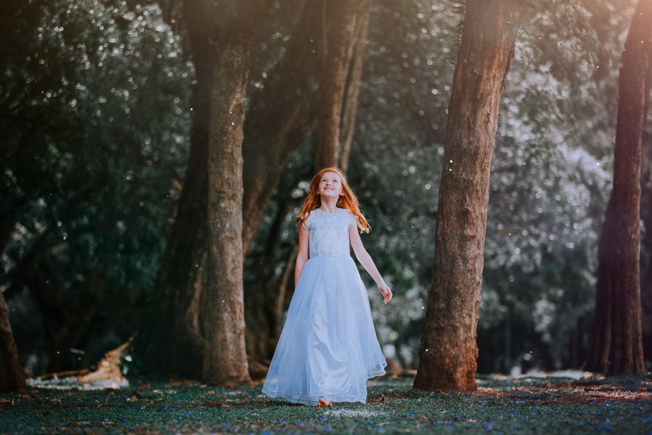A young girl in a flowing blue dress joyfully walks through a sunlit forest.