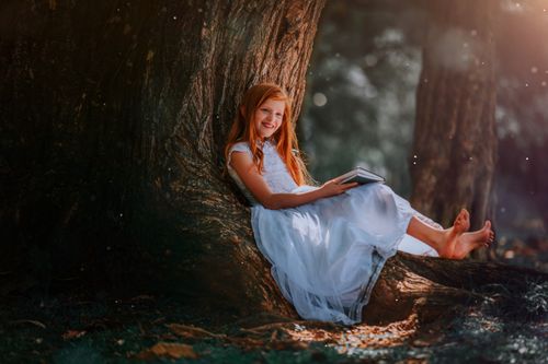Free A joyful girl in a white dress enjoys reading under a big tree on a sunny day. Stock Photo