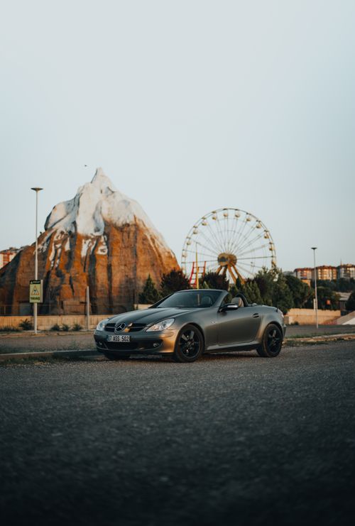 Free Luxury convertible car parked near an amusement park with a ferris wheel and mountain backdrop. Stock Photo
