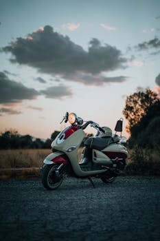 A vintage motor scooter parked on a gravel path at sunset under a cloudy sky.