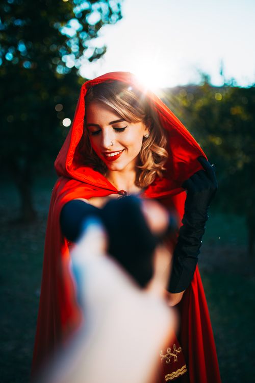 Free Portrait of a woman wearing a red cape, smiling brightly in a sunlit forest. Stock Photo