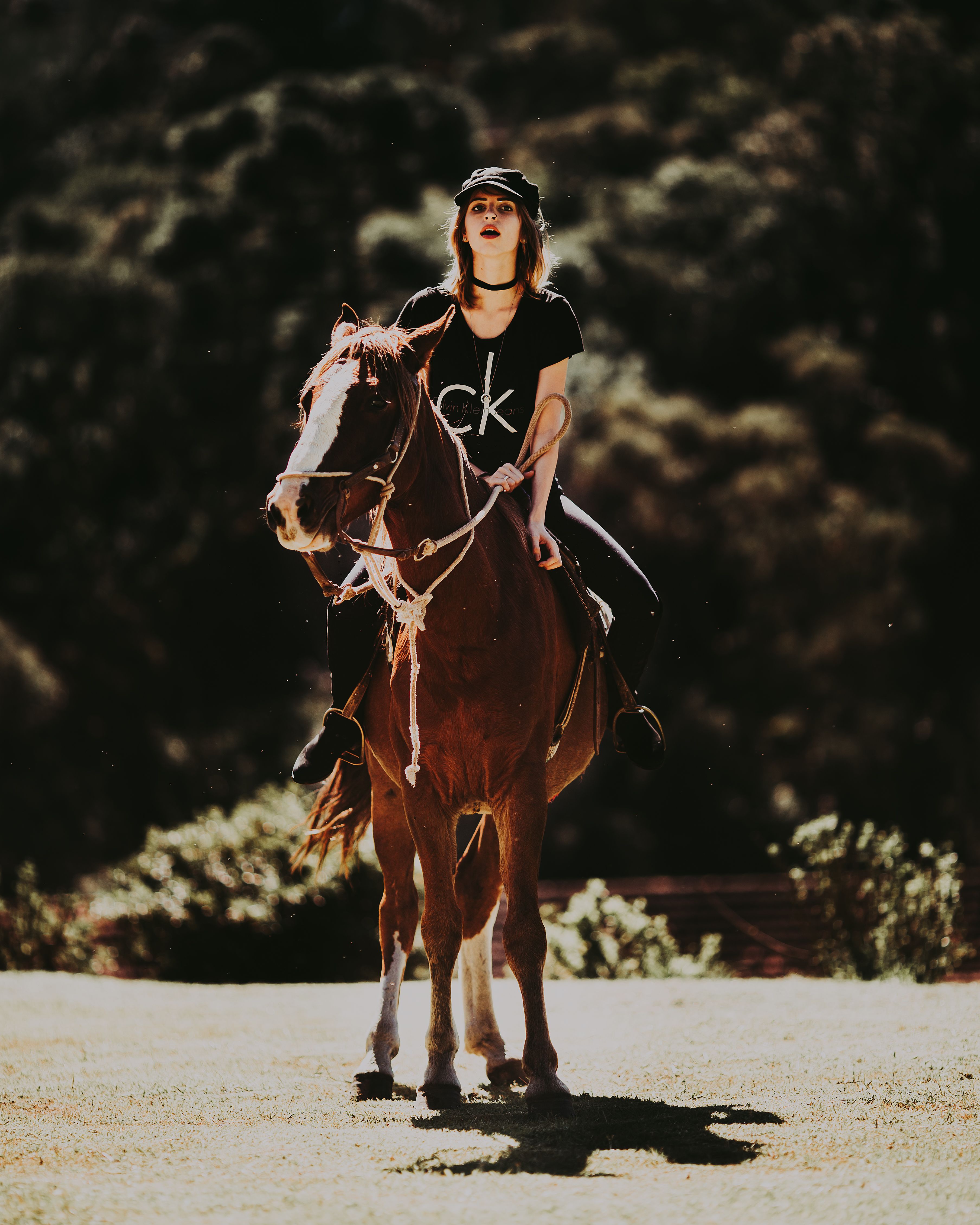 A young woman in casual attire rides a horse through a sunny outdoor setting.