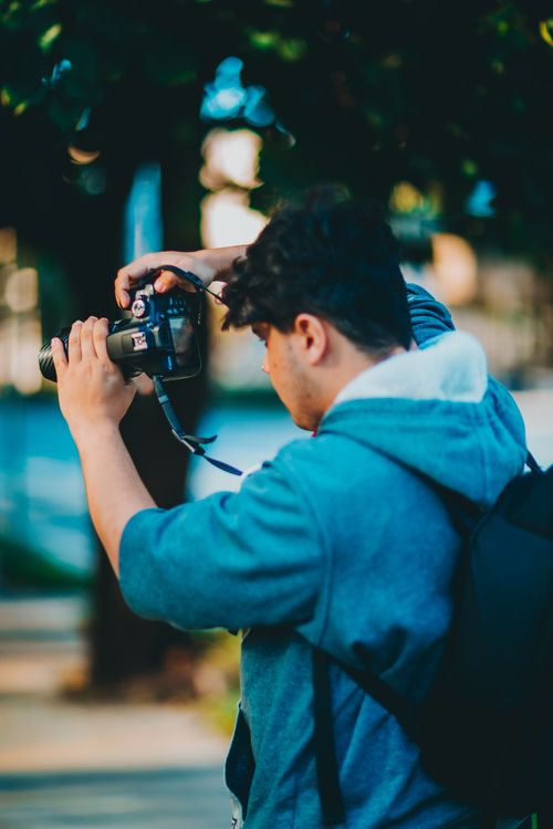 Free A young man in a blue jacket uses a DSLR camera outdoors, capturing a lively scene. Stock Photo