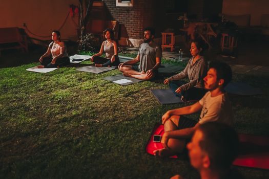 A diverse group meditating outdoors on yoga mats during evening time.