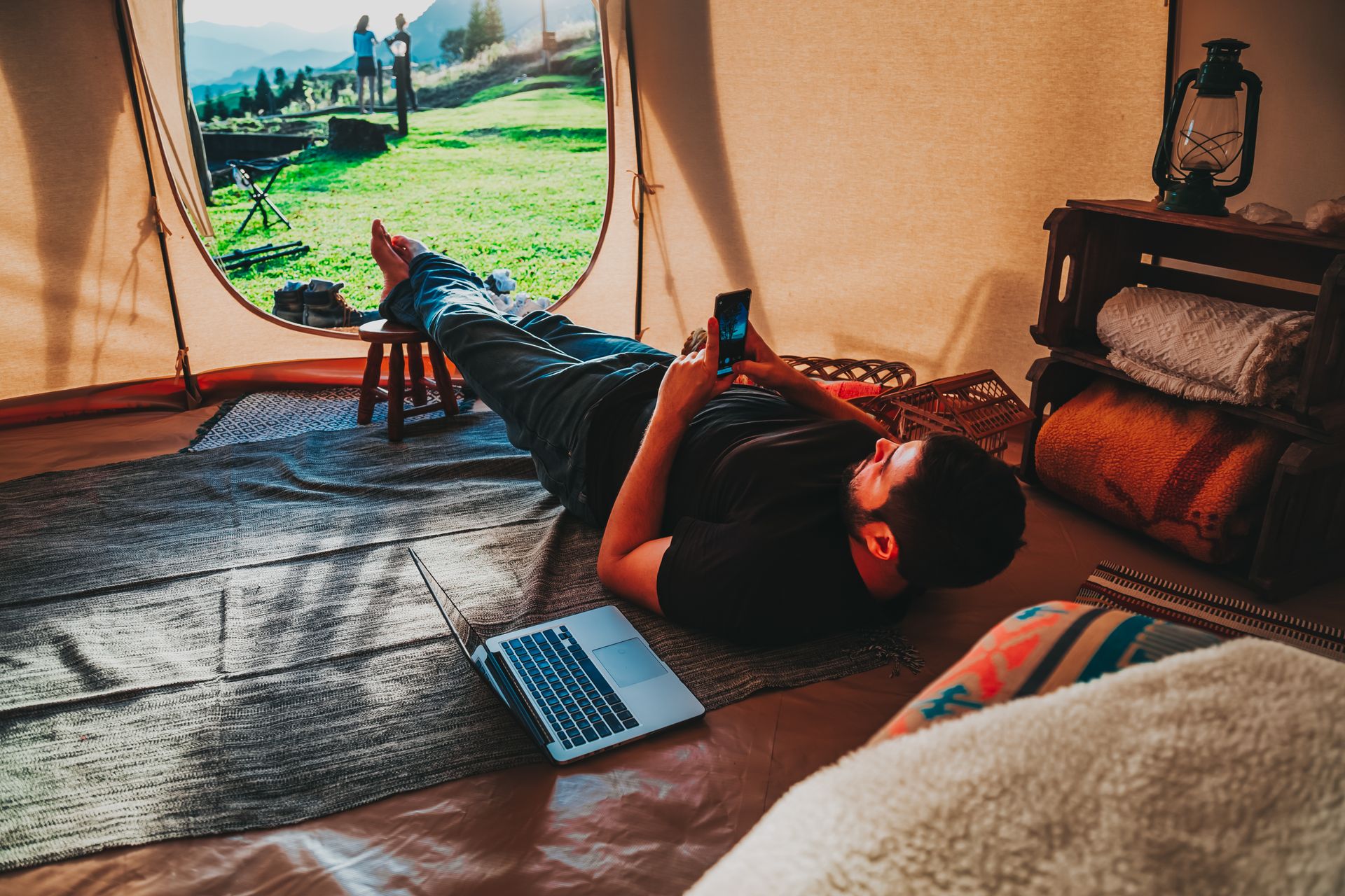 Man relaxing in a tent using smartphone and laptop, depicting a mix of nature and technology.