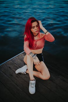 Young woman with dyed red hair posing on a wooden jetty by the sea, embodying youthful fashion and style.