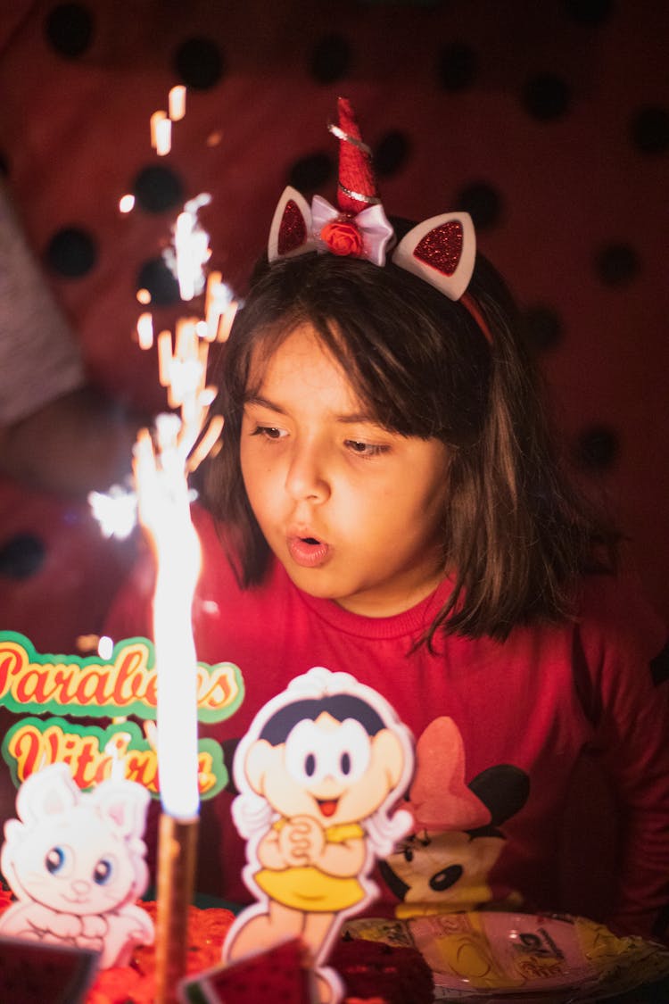 A Girl In Pink Sweater Wearing Headband While Blowing The Sparkler Candle