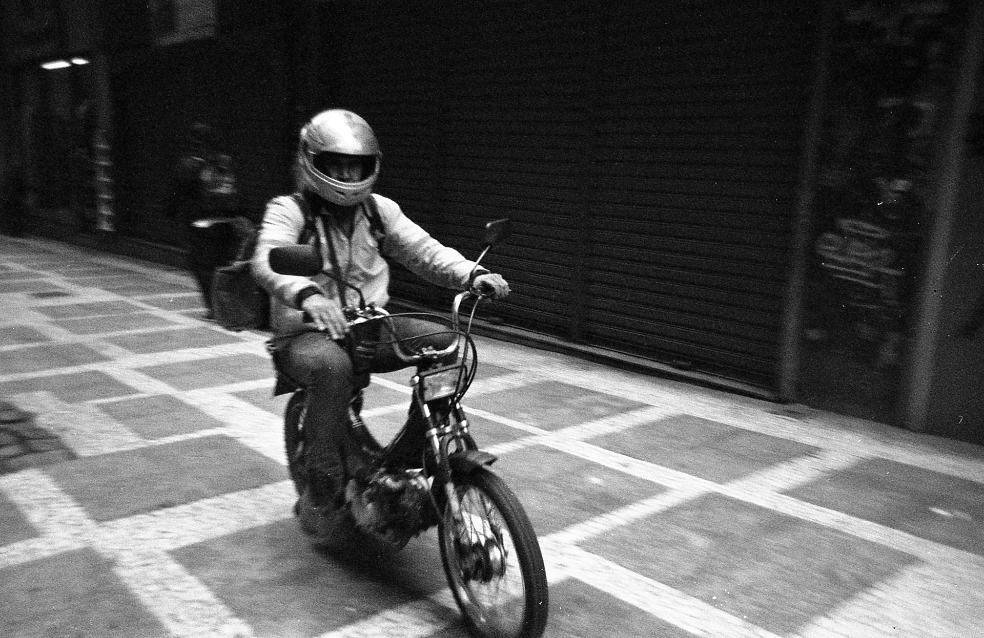 Black and white photo of a motorcyclist riding on a city street with a closed shop in the background.
