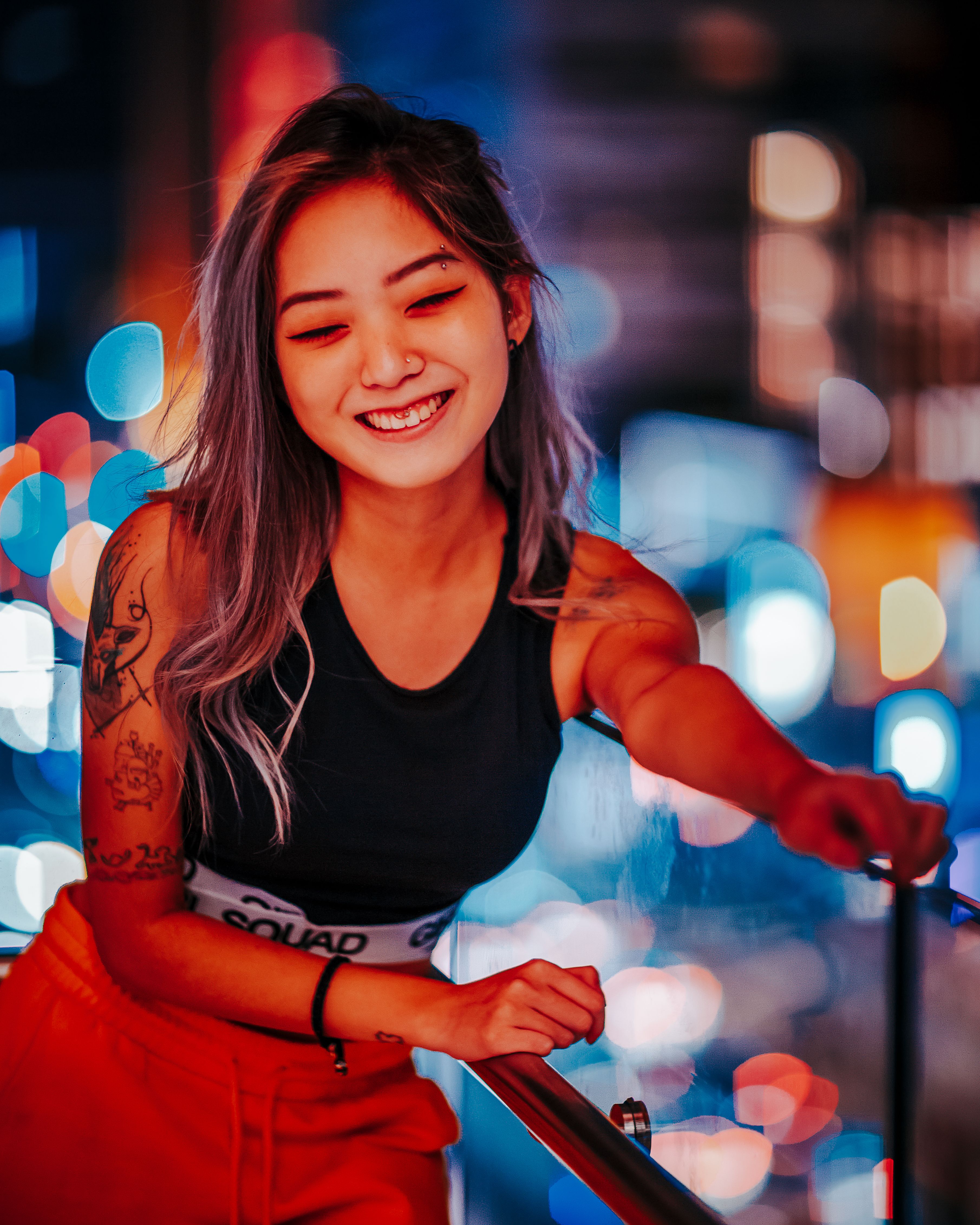 A joyful tattooed woman with long hair smiles under city lights, creating a vibrant and warm portrait.