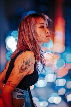 Portrait of a young woman with tattoos looking over a cityscape at night with vibrant bokeh lights.