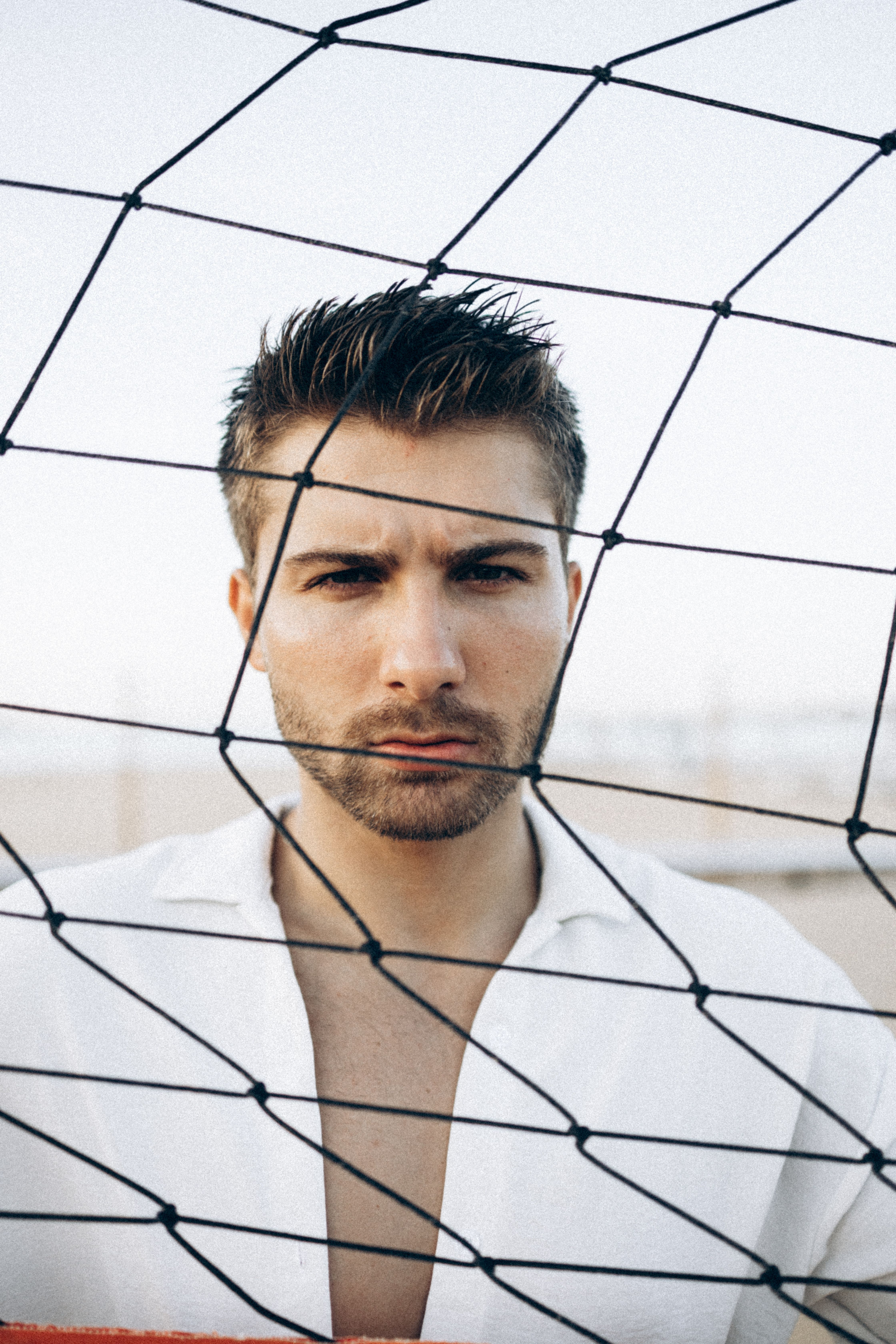Close-up of a serious man behind a net, wearing a white shirt, outdoors during daytime.