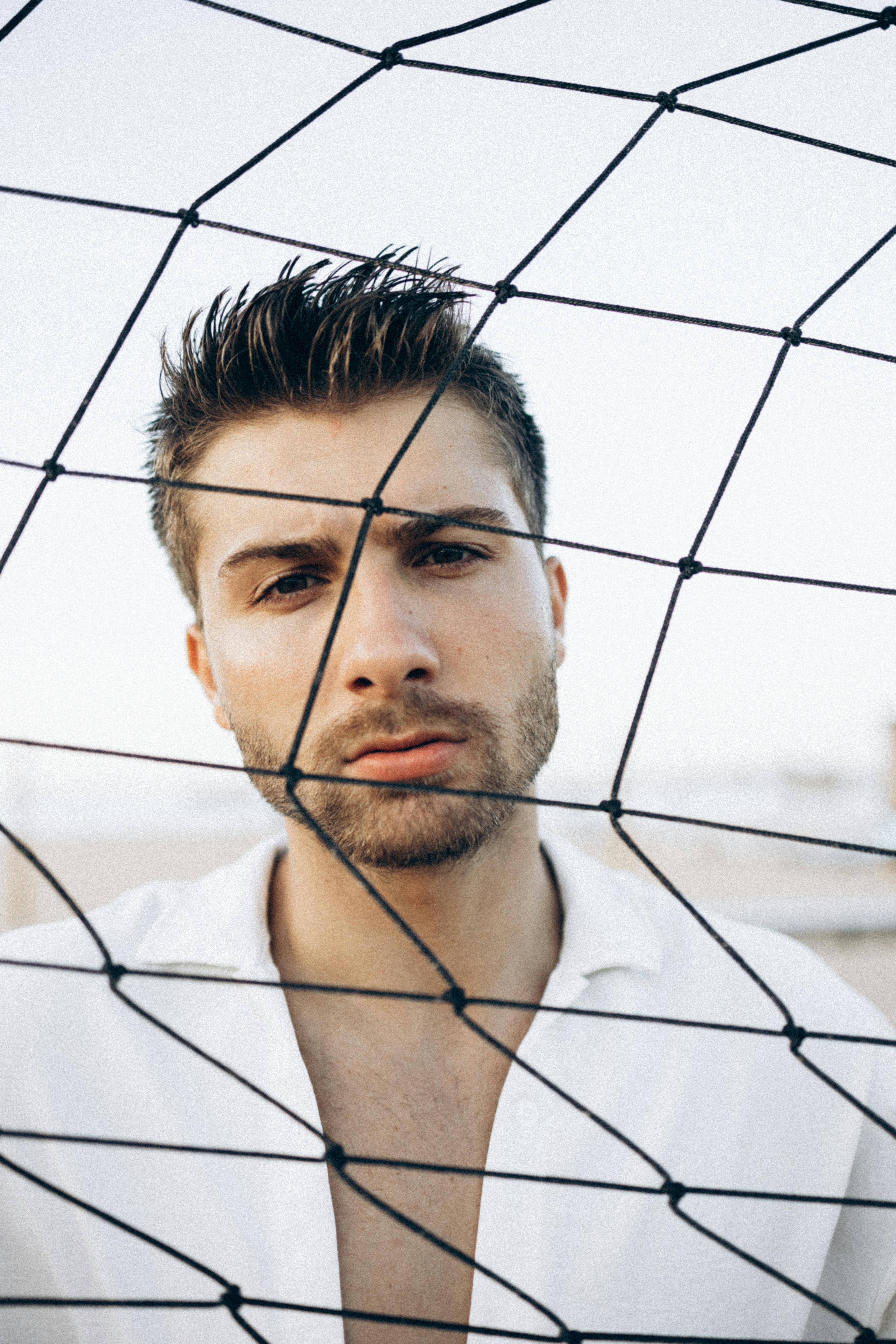 Close-up portrait of a bearded man in a white shirt behind netting with a pensive expression.