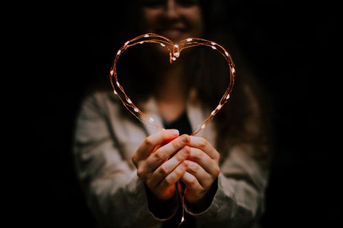 Free A woman holding heart-shaped string lights against a dark blurred background, creating a warm, intimate mood. Stock Photo