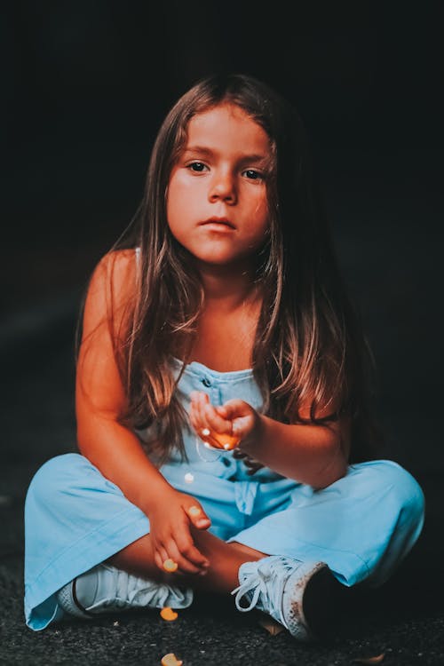 Photo of a Girl with Long Hair Wearing Blue Dungarees Sitting on a Ground and Holding Glitter