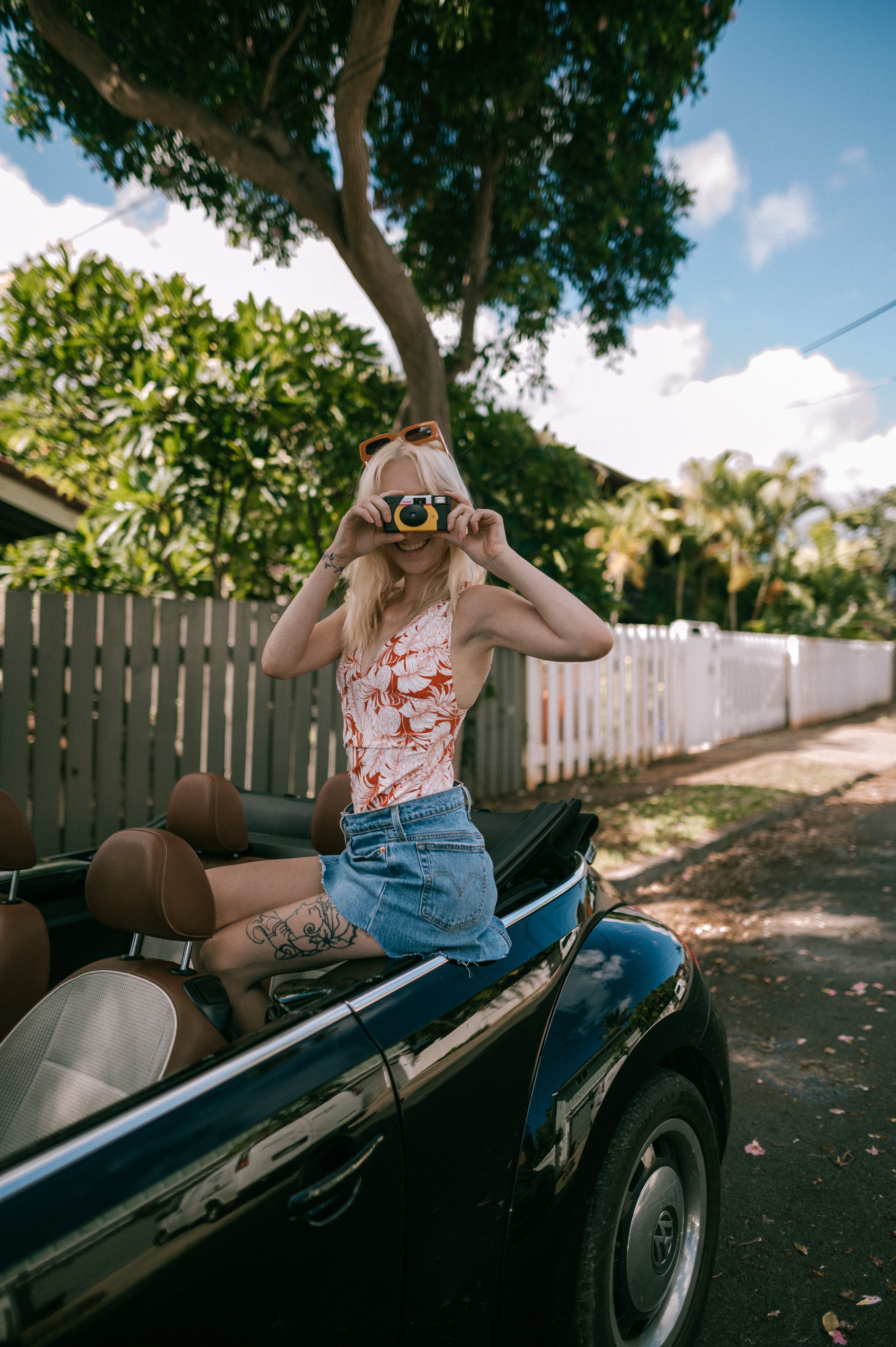 Young woman in a convertible capturing a moment with her camera on a sunny city street.