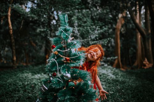 Free A joyful red-haired child playing hide and seek behind a decorated Christmas tree in a forest setting. Stock Photo