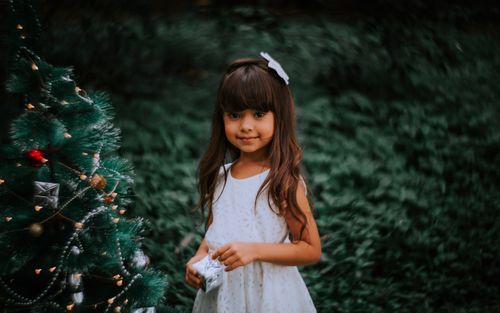 Free A young girl in a white dress stands by a decorated Christmas tree outdoors, holding a gift. Stock Photo