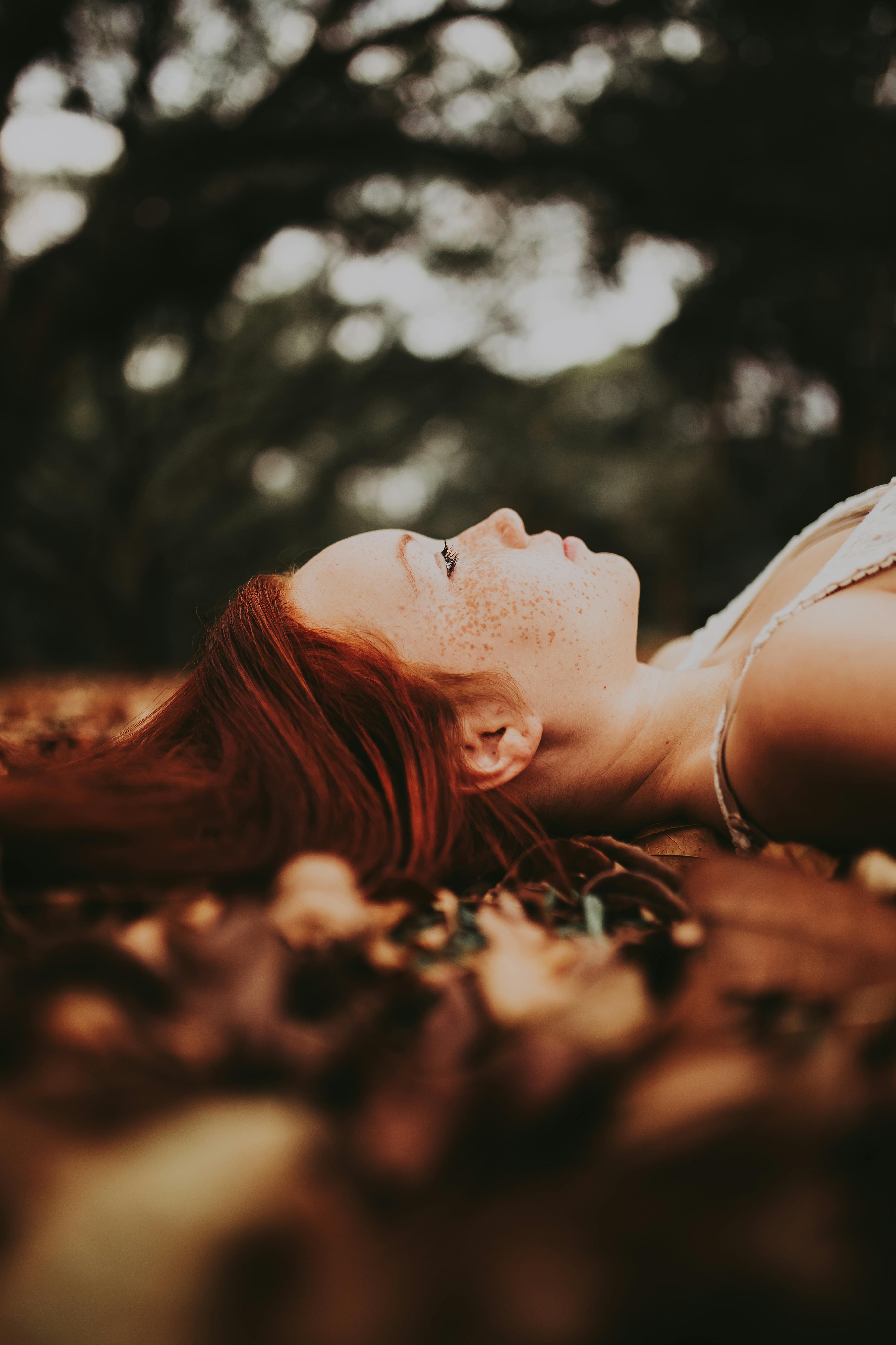 A young redhead woman peacefully lies on autumn leaves in a serene forest setting.