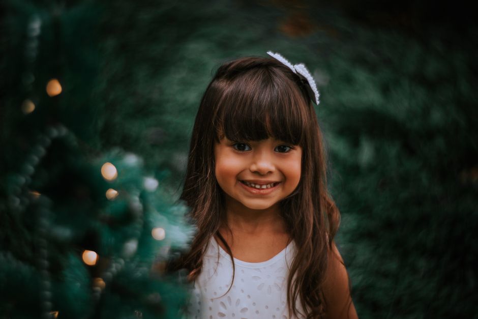 A cheerful young girl with bangs smiles near a decorated Christmas tree.