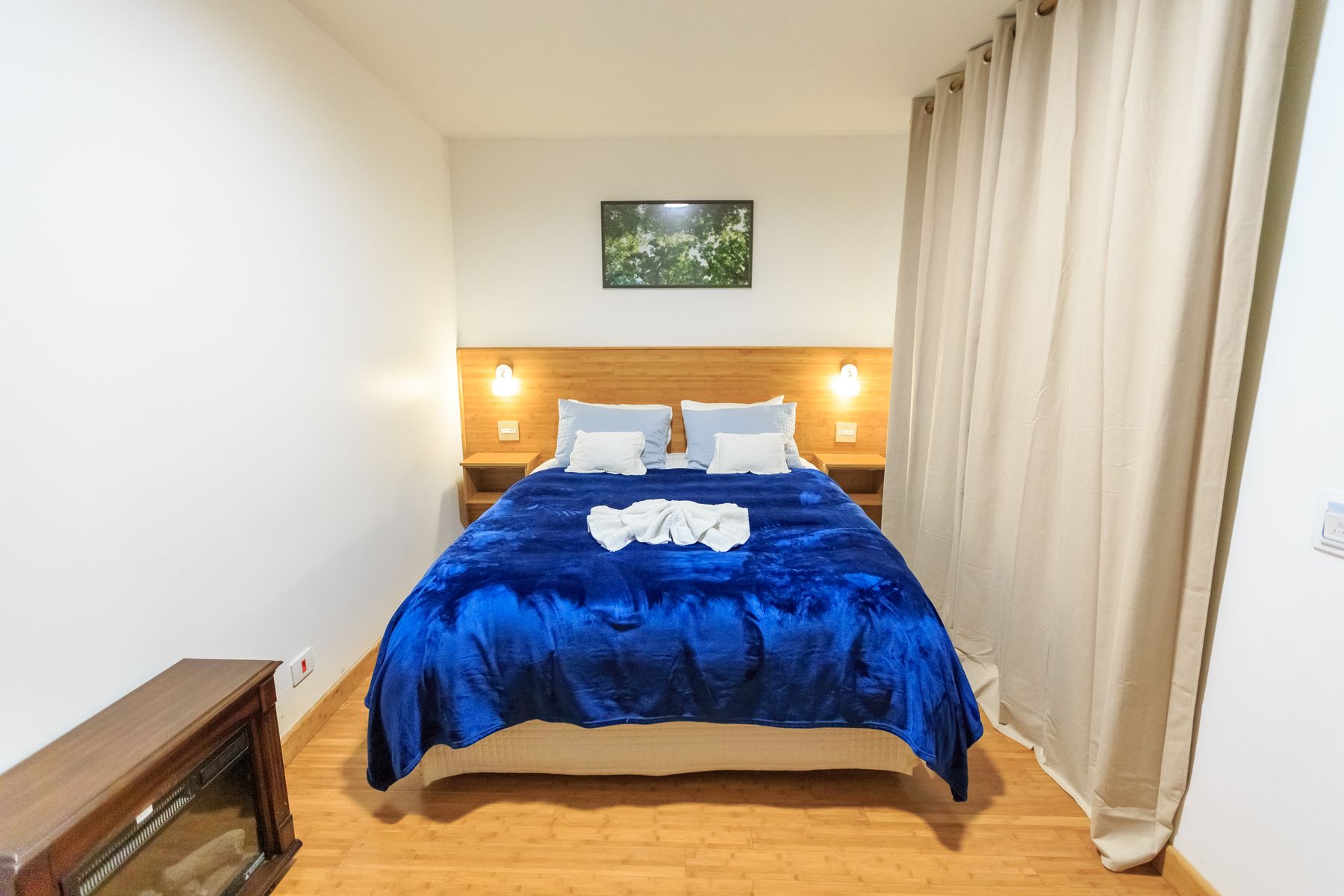 Compact hotel bedroom with a deep blue velvet bed, blue walls, pale headboard and warm bedside lights
