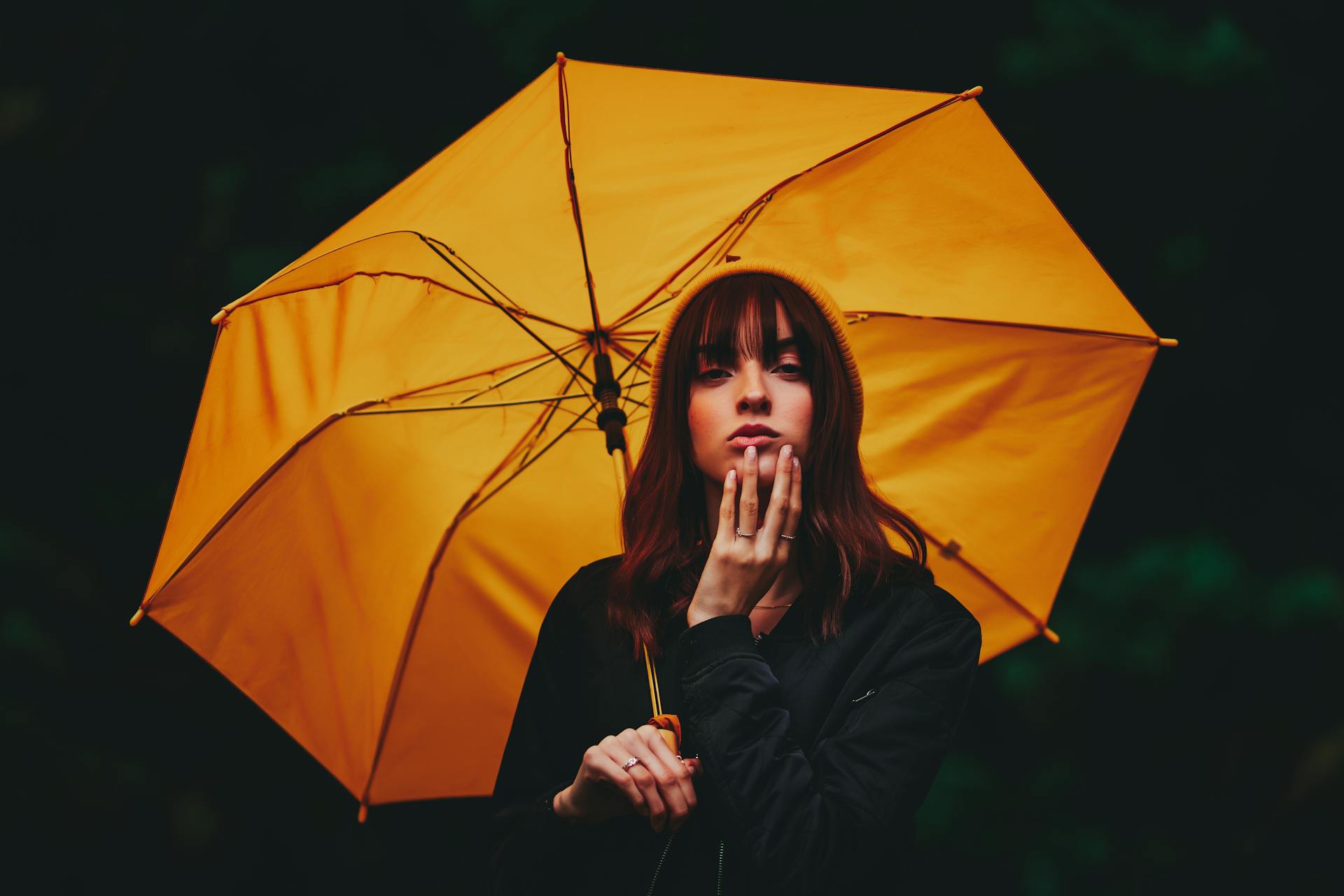 A woman poses thoughtfully with a vibrant yellow umbrella against a dark background.